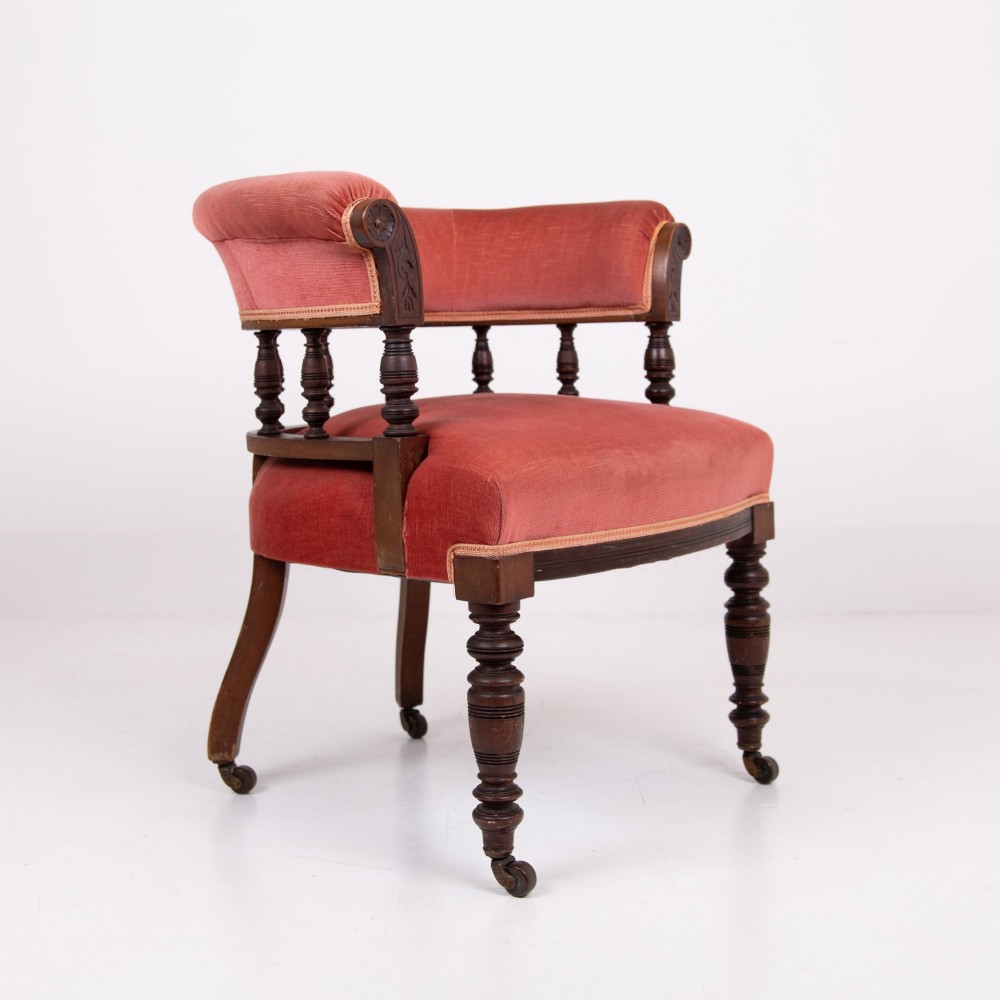 A late Victorian armchair featuring a curved horseshoe back rest, turned wood spindles, terracotta fabric upholstery, and turned front legs on castors. View V