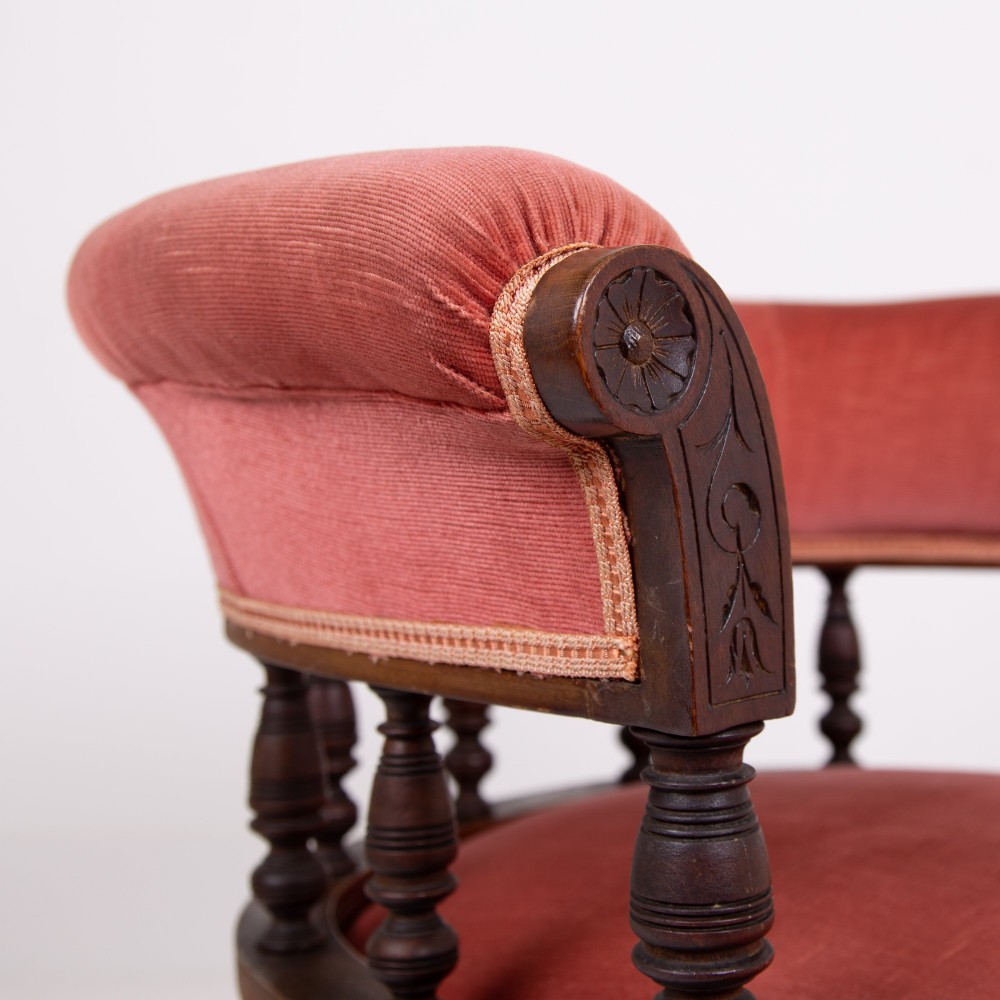 A late Victorian armchair featuring a curved horseshoe back rest, turned wood spindles, terracotta fabric upholstery, and turned front legs on castors. View VI