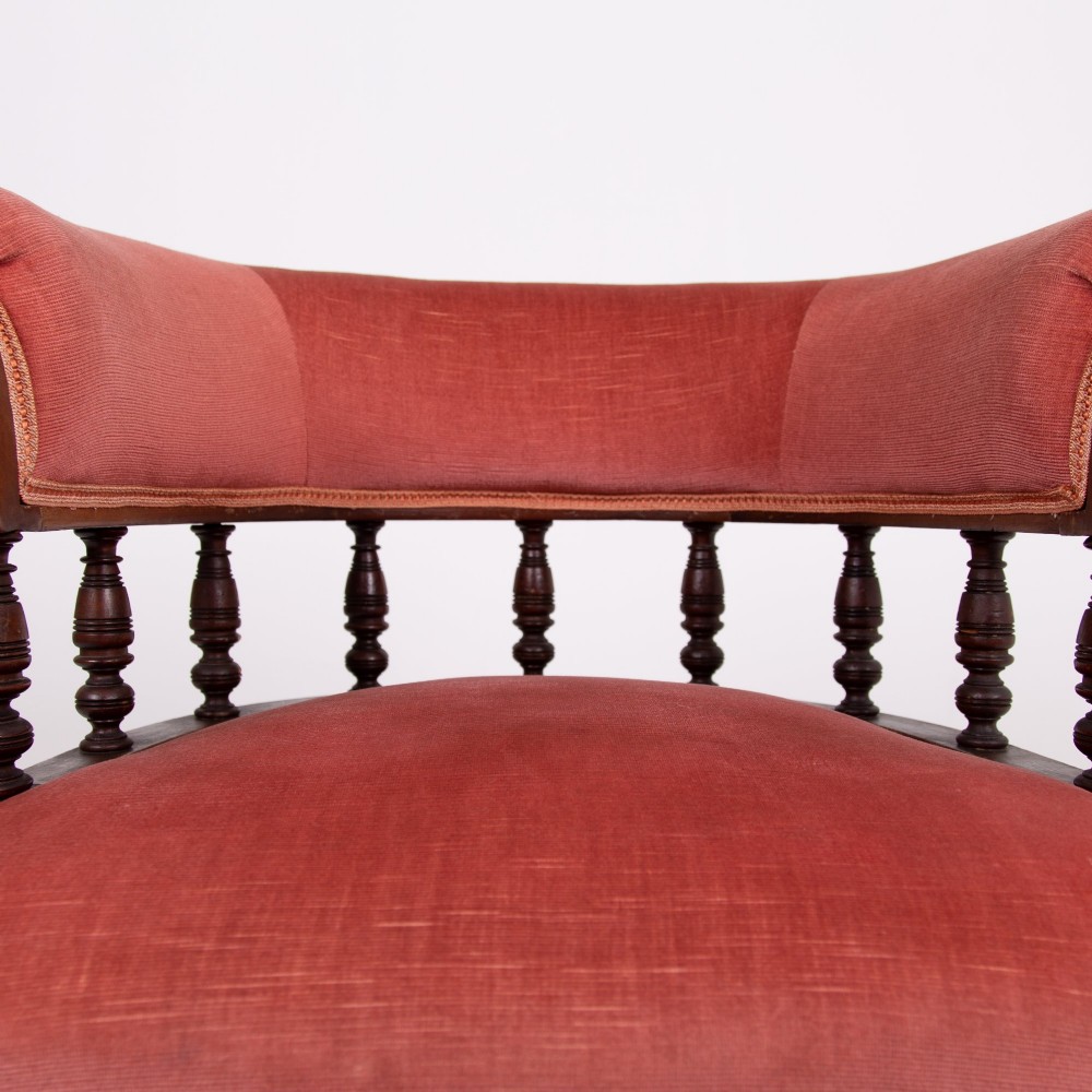 A late Victorian armchair featuring a curved horseshoe back rest, turned wood spindles, terracotta fabric upholstery, and turned front legs on castors. View VII