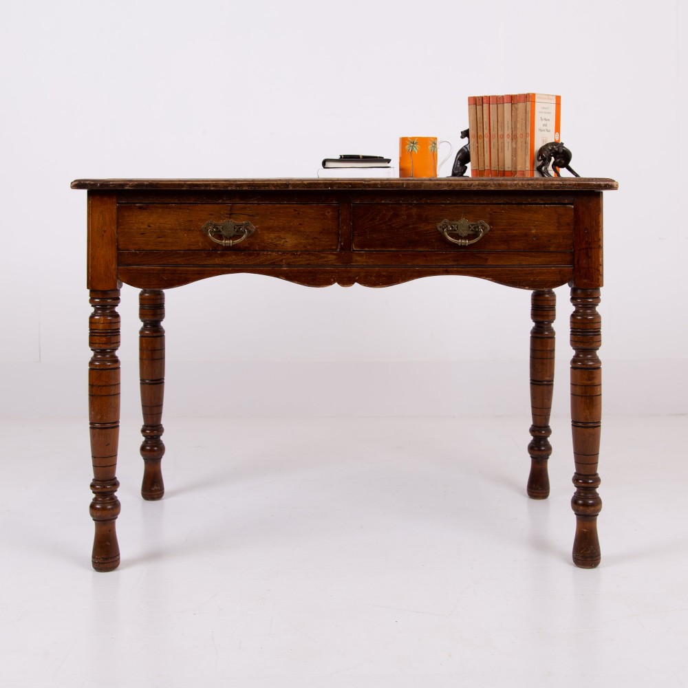 A brown walnut writing desk featuring two frieze drawers with brass handles, turned legs, and a rectangular inset leather top.