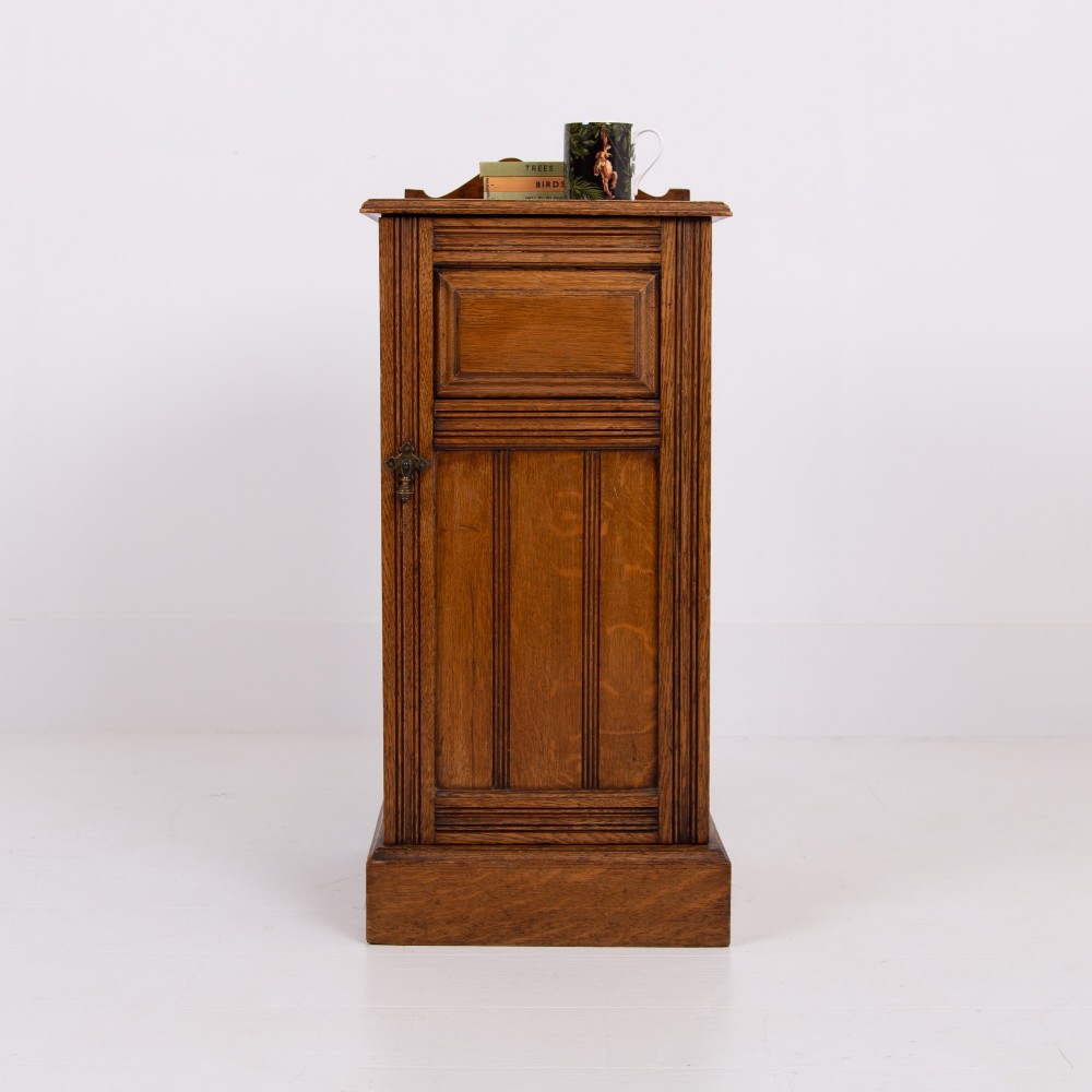 A slim Victorian oak pot cupboard featuring a single panelled door with a small brass latch, resting on a solid plinth base with a gallery top.