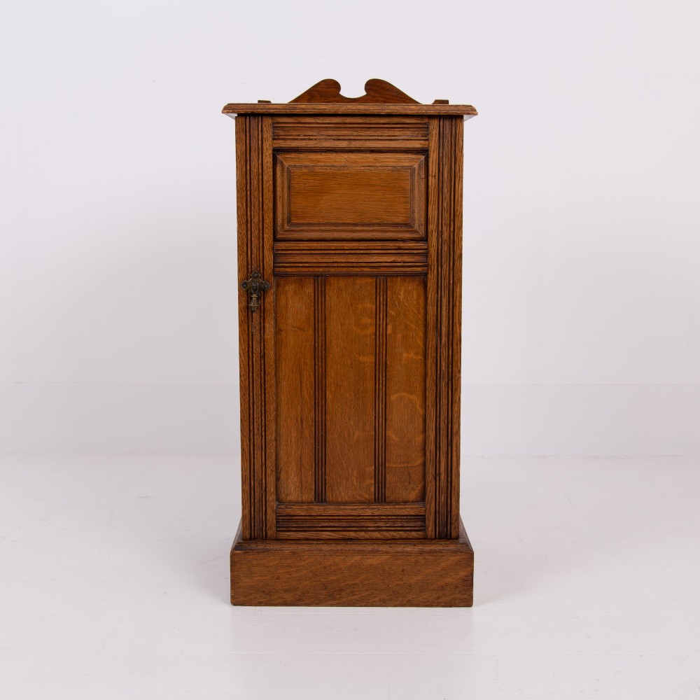 A slim Victorian oak pot cupboard featuring a single panelled door with a small brass latch, resting on a solid plinth base with a gallery top. View II