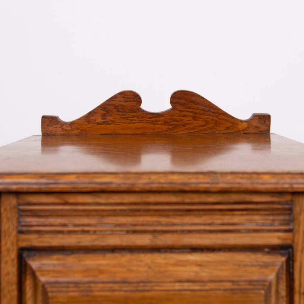 A slim Victorian oak pot cupboard featuring a single panelled door with a small brass latch, resting on a solid plinth base with a gallery top. View IV