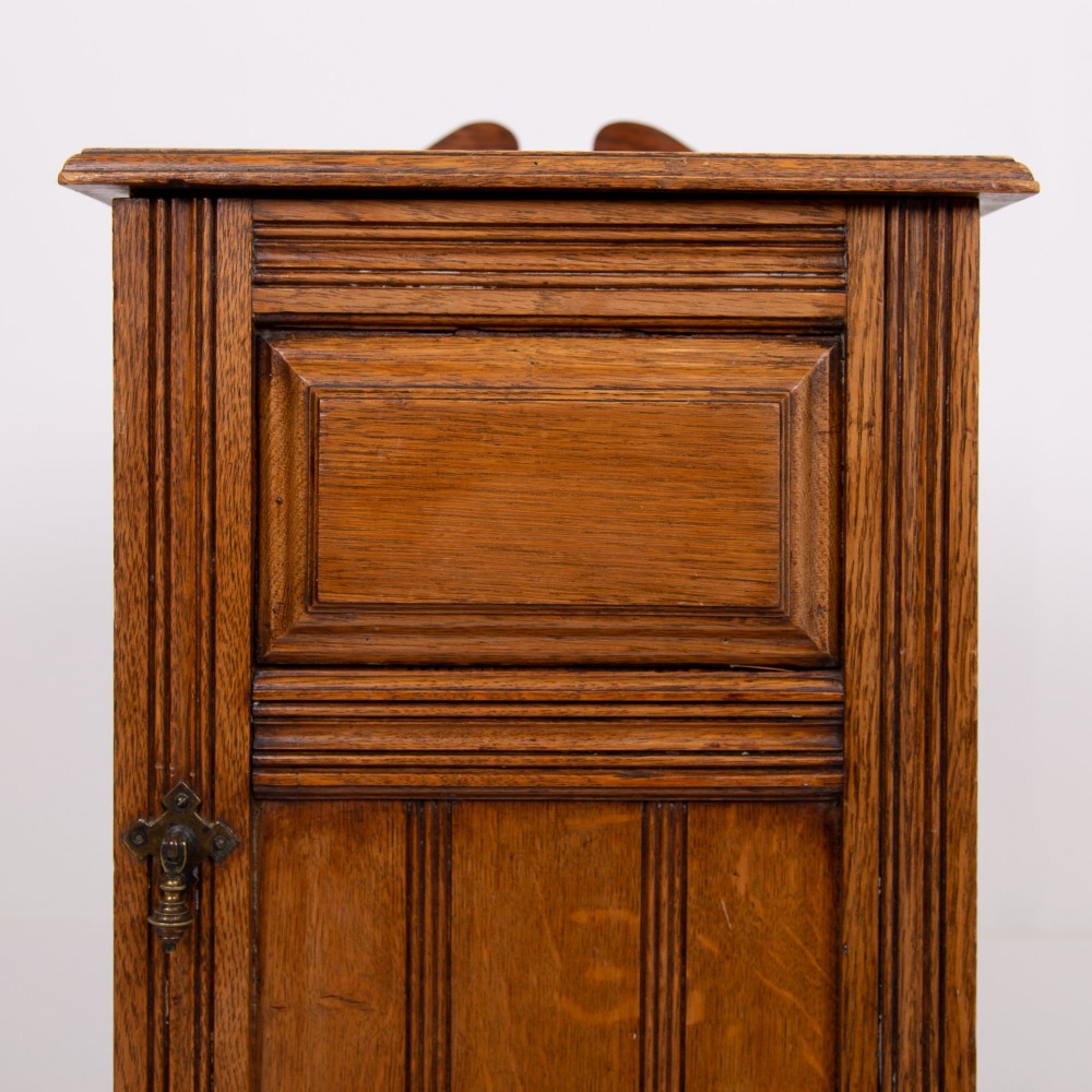 A slim Victorian oak pot cupboard featuring a single panelled door with a small brass latch, resting on a solid plinth base with a gallery top. View V