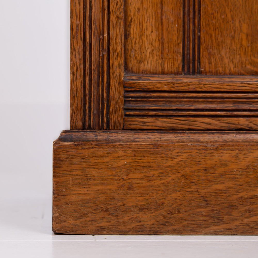 A slim Victorian oak pot cupboard featuring a single panelled door with a small brass latch, resting on a solid plinth base with a gallery top. View VI