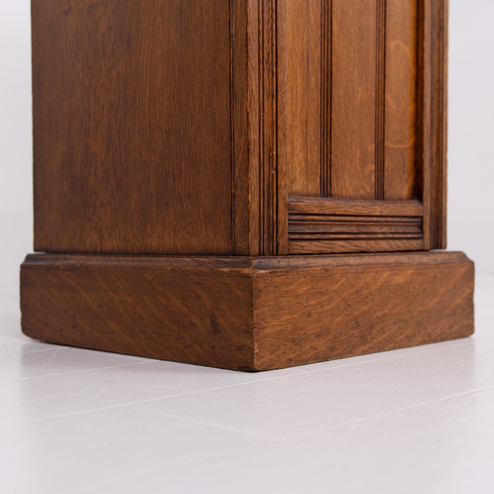 A slim Victorian oak pot cupboard featuring a single panelled door with a small brass latch, resting on a solid plinth base with a gallery top. View VII