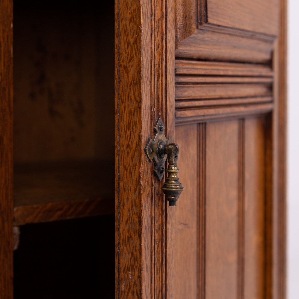 A slim Victorian oak pot cupboard featuring a single panelled door with a small brass latch, resting on a solid plinth base with a gallery top. View IX