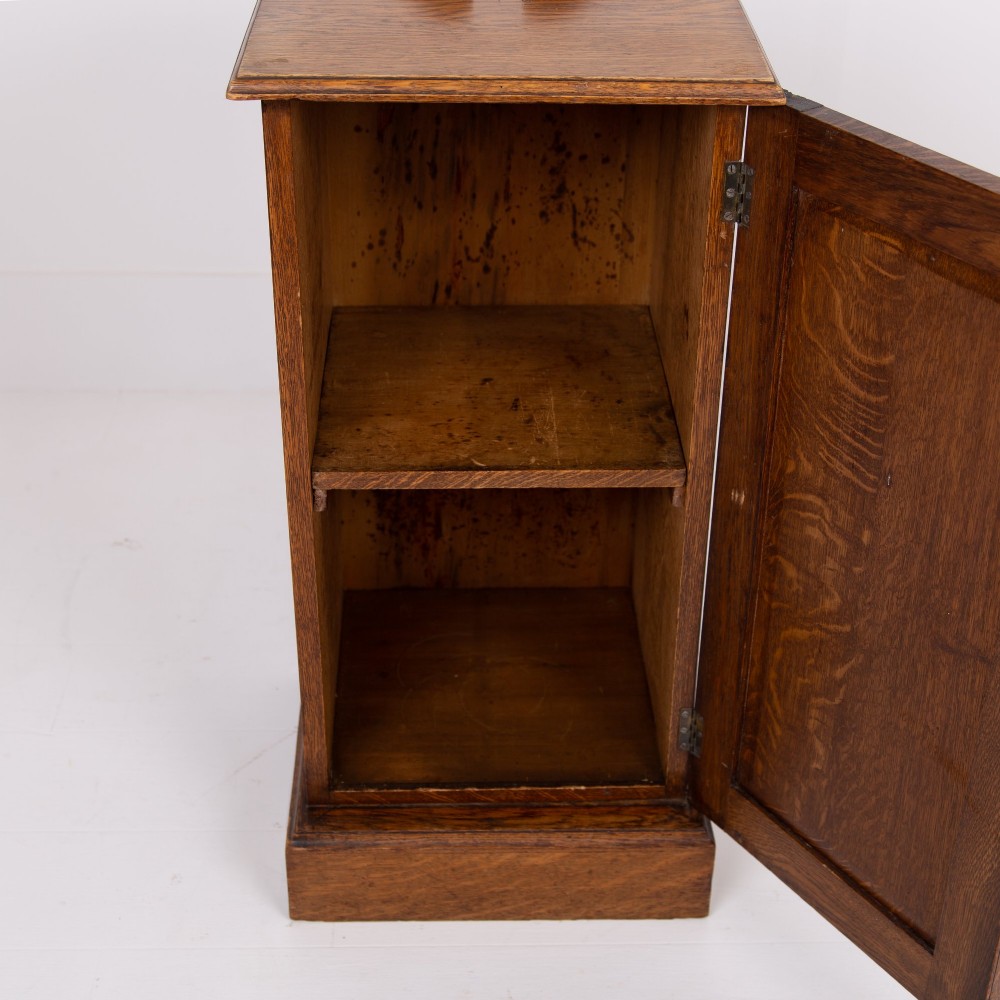 A slim Victorian oak pot cupboard featuring a single panelled door with a small brass latch, resting on a solid plinth base with a gallery top. View X