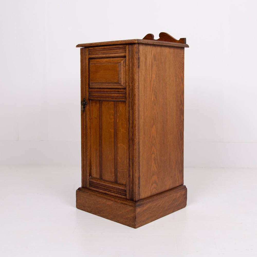 A slim Victorian oak pot cupboard featuring a single panelled door with a small brass latch, resting on a solid plinth base with a gallery top. View XI