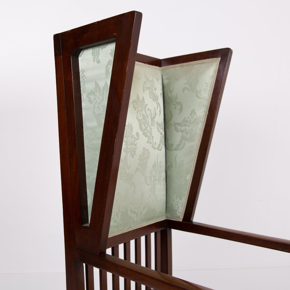 An Arts and Crafts high-backed armchair crafted from dark timber with vertical under-arm slats, architectural wings, and pale green patterned fabric upholstery. View VI