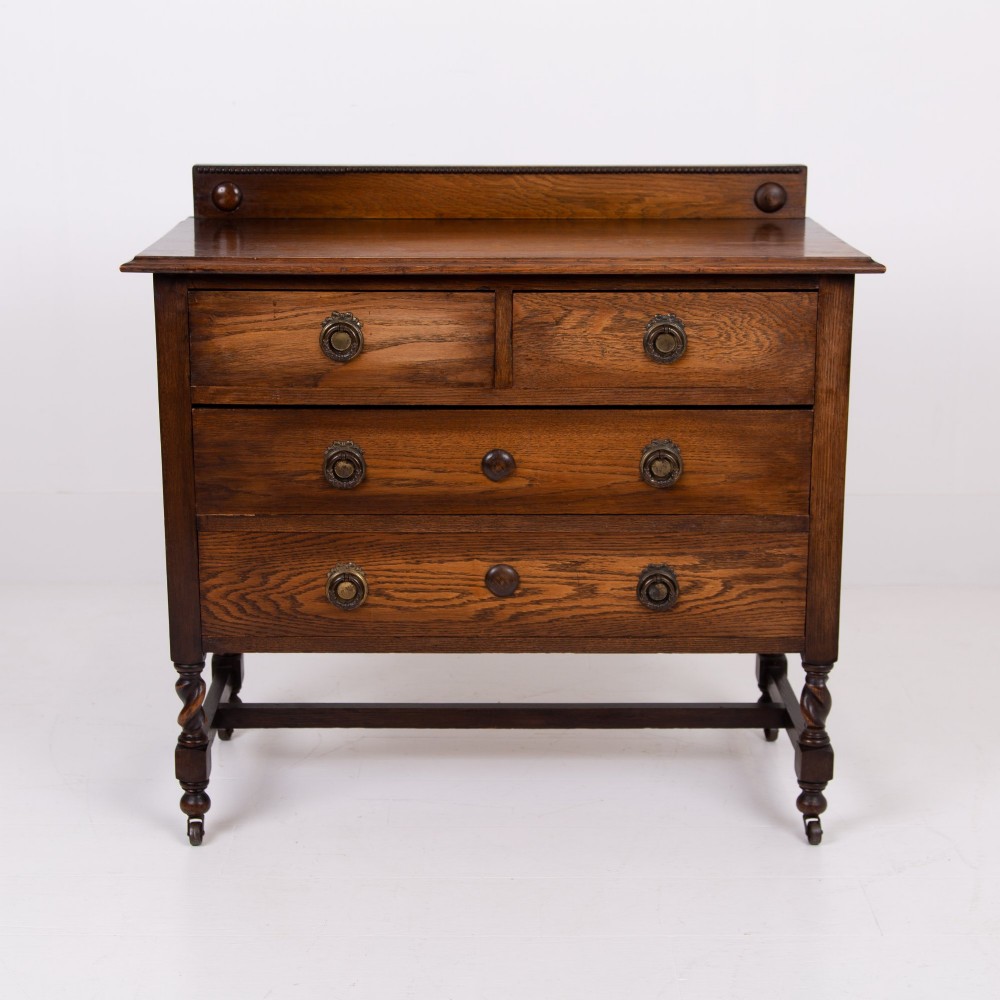 An Edwardian oak chest of drawers with two short over two long drawers, brass drop handles, a low backstand, and barley twist legs set on castors. View II
