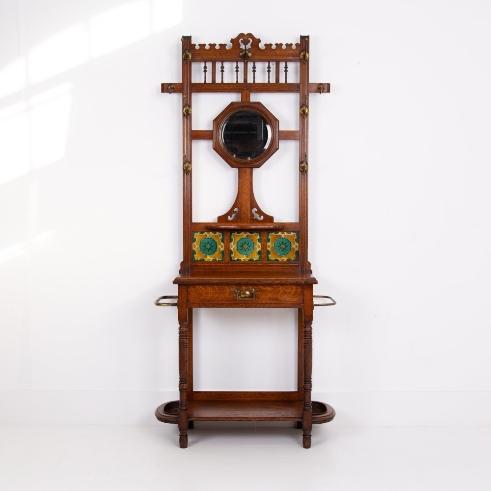A tall Victorian golden oak hallstand featuring an octagonal central mirror, decorative ceramic tiles, brass coat hooks, a single central drawer, and lower umbrella drip trays.