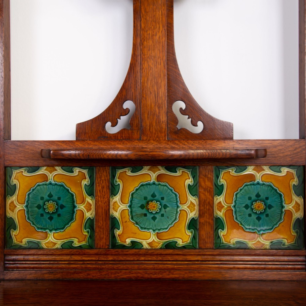 A tall Victorian golden oak hallstand featuring an octagonal central mirror, decorative ceramic tiles, brass coat hooks, a single central drawer, and lower umbrella drip trays. View II
