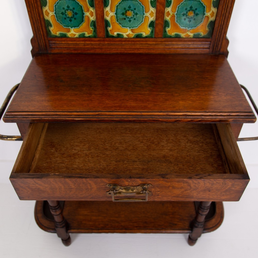 A tall Victorian golden oak hallstand featuring an octagonal central mirror, decorative ceramic tiles, brass coat hooks, a single central drawer, and lower umbrella drip trays. View VII