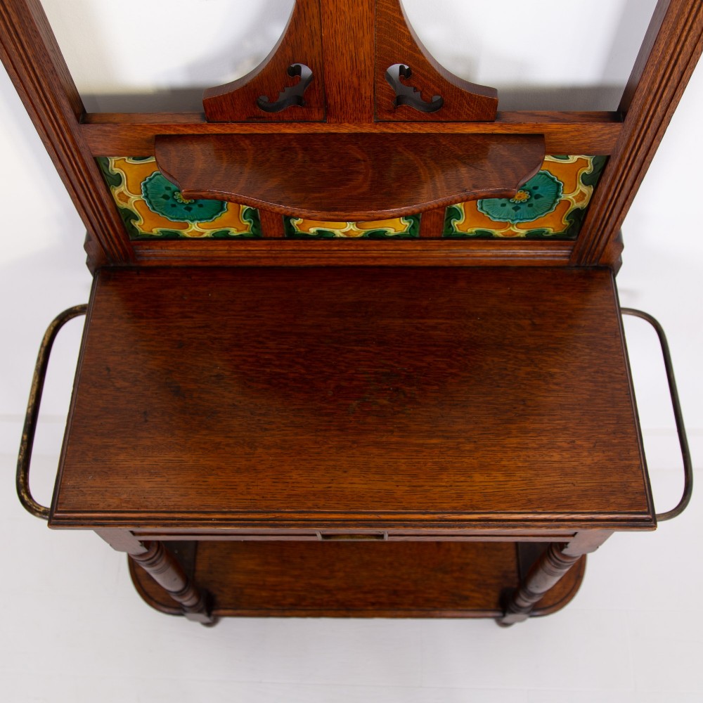 A tall Victorian golden oak hallstand featuring an octagonal central mirror, decorative ceramic tiles, brass coat hooks, a single central drawer, and lower umbrella drip trays. View IX