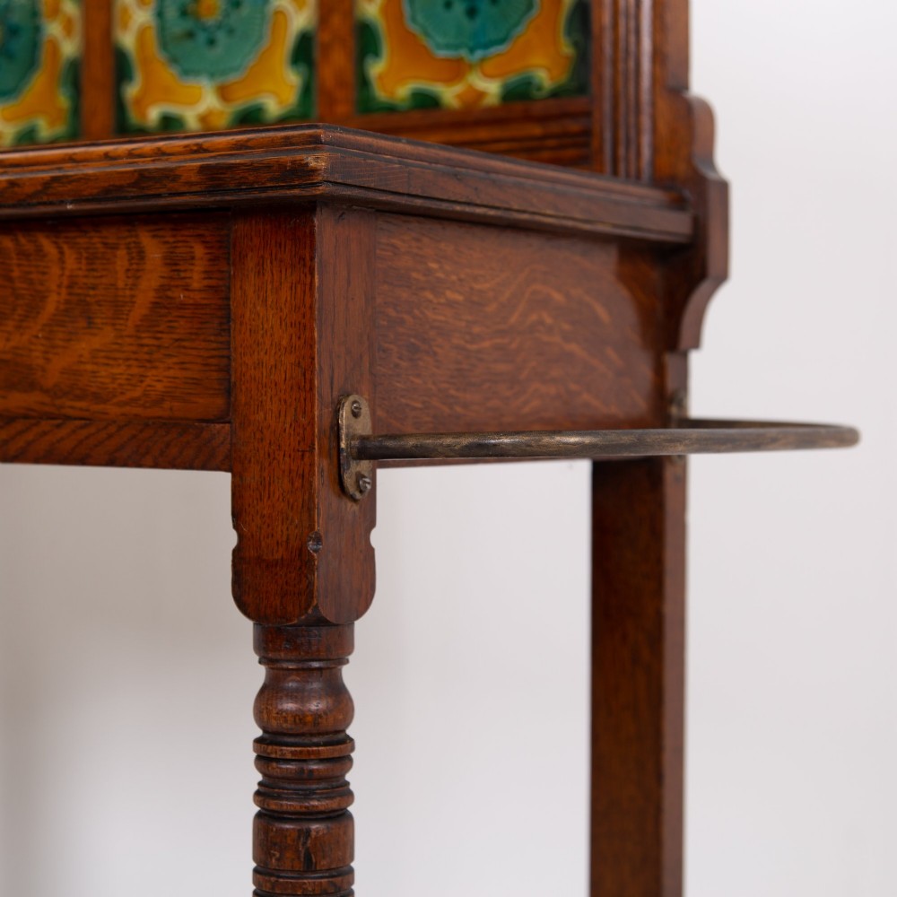 A tall Victorian golden oak hallstand featuring an octagonal central mirror, decorative ceramic tiles, brass coat hooks, a single central drawer, and lower umbrella drip trays. View XII