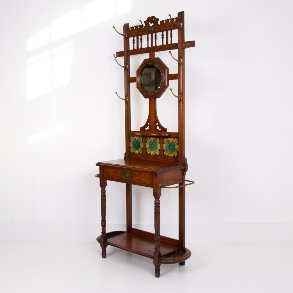 A tall Victorian golden oak hallstand featuring an octagonal central mirror, decorative ceramic tiles, brass coat hooks, a single central drawer, and lower umbrella drip trays. View XIV