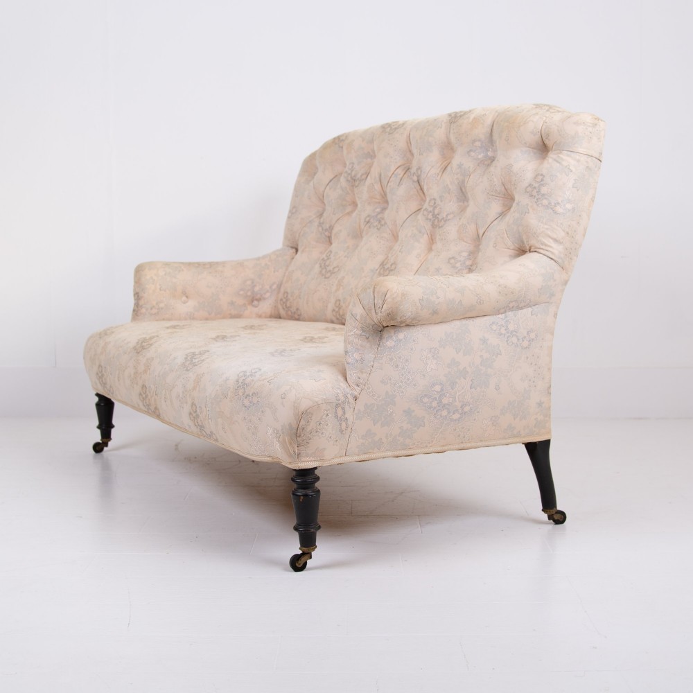 An antique French Napoleon III sofa upholstered in pale cream patterned fabric with deep buttoning, curved backrest, and turned dark wooden legs on brass castors. View II