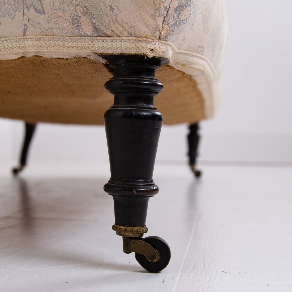 An antique French Napoleon III sofa upholstered in pale cream patterned fabric with deep buttoning, curved backrest, and turned dark wooden legs on brass castors. View X