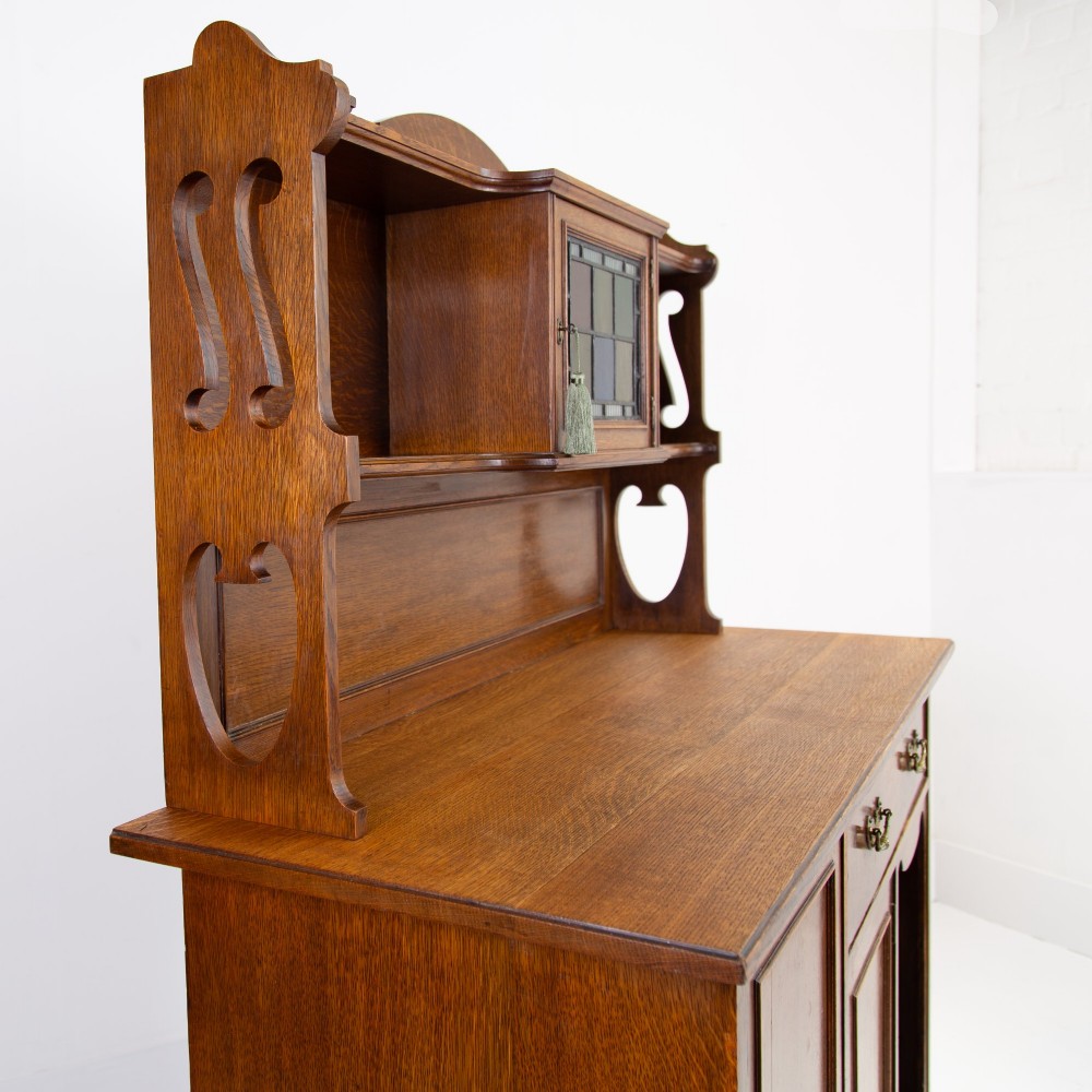 An Arts and Crafts oak cabinet dresser featuring an upper shelf unit with a central stained glass cabinet door, side heart cutouts, lower cupboards, a single drawer, and an open side niche. View X