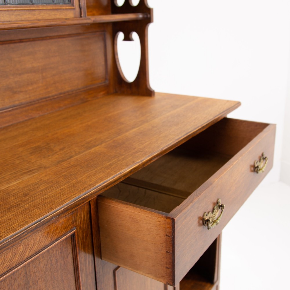 An Arts and Crafts oak cabinet dresser featuring an upper shelf unit with a central stained glass cabinet door, side heart cutouts, lower cupboards, a single drawer, and an open side niche. View XI