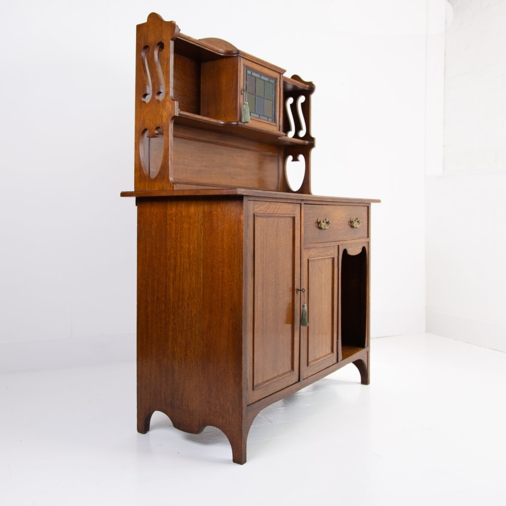 An Arts and Crafts oak cabinet dresser featuring an upper shelf unit with a central stained glass cabinet door, side heart cutouts, lower cupboards, a single drawer, and an open side niche. View XII