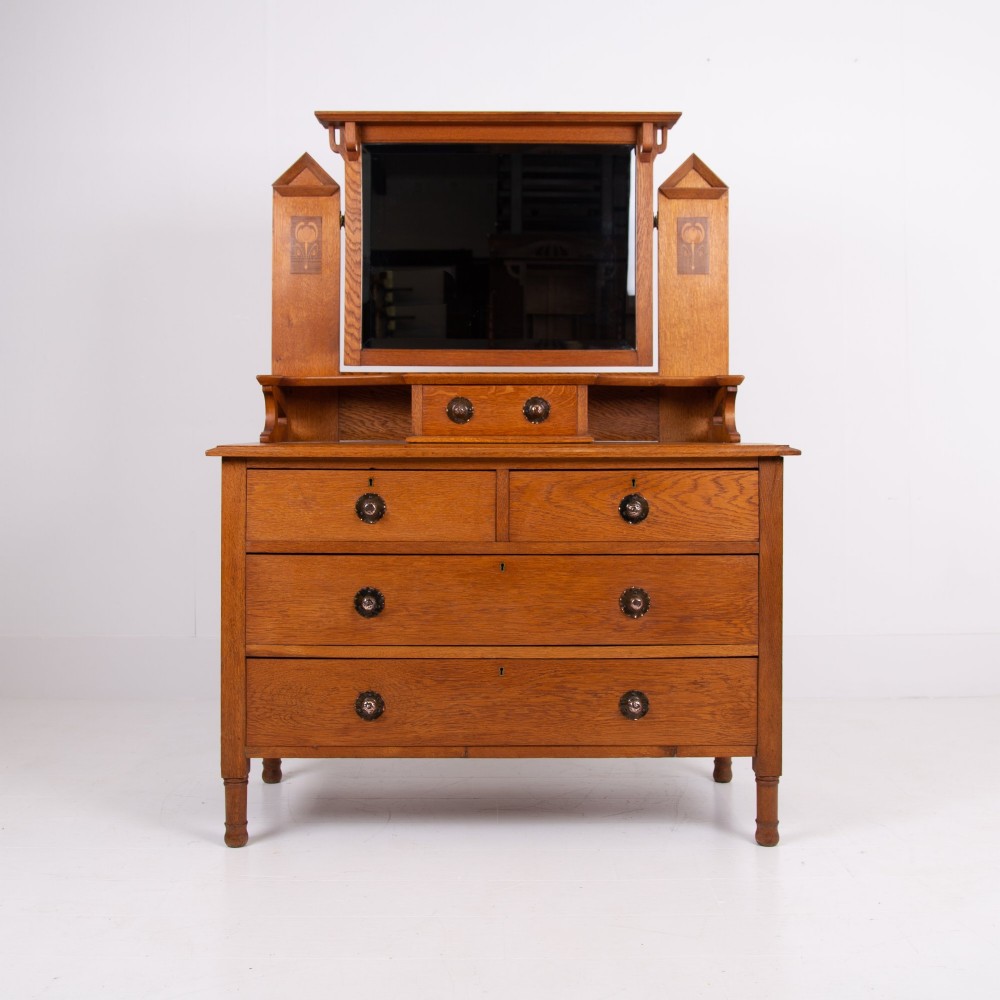 An Arts and Crafts oak dressing chest featuring a superstructure with a central mirror, inlaid side panels, and small jewellery drawers above a chest of drawers. View II
