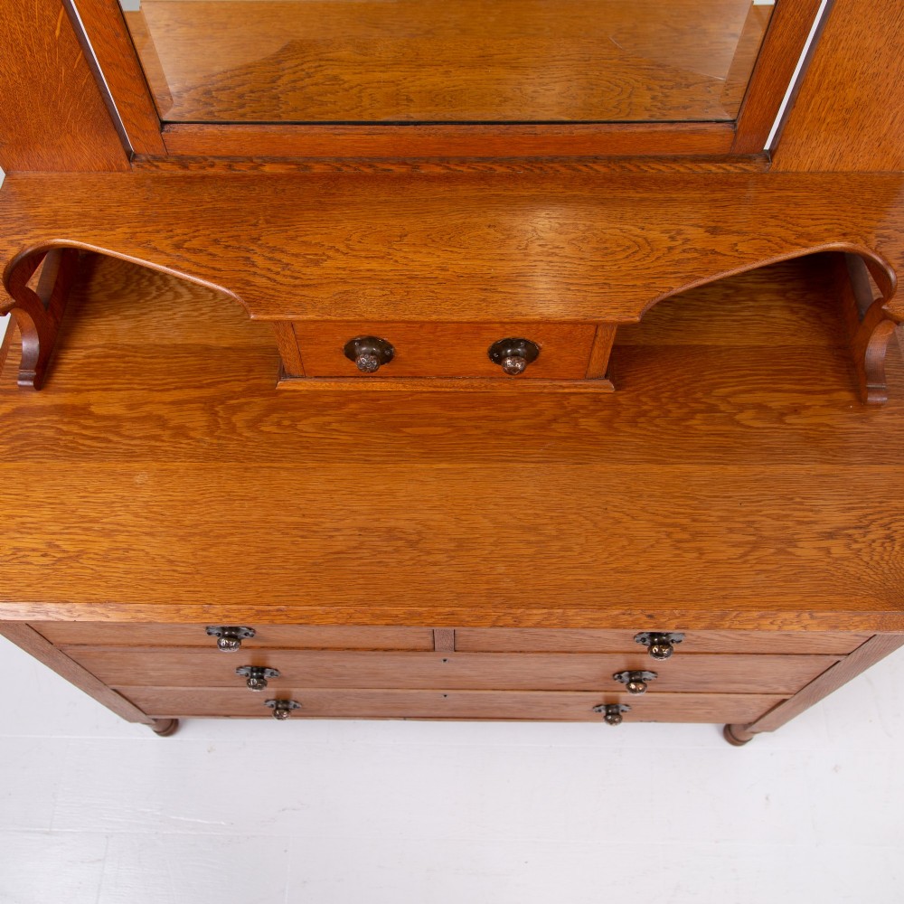 An Arts and Crafts oak dressing chest featuring a superstructure with a central mirror, inlaid side panels, and small jewellery drawers above a chest of drawers. View X