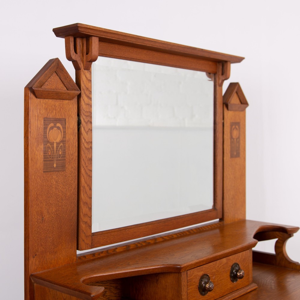 An Arts and Crafts oak dressing chest featuring a superstructure with a central mirror, inlaid side panels, and small jewellery drawers above a chest of drawers. View XI