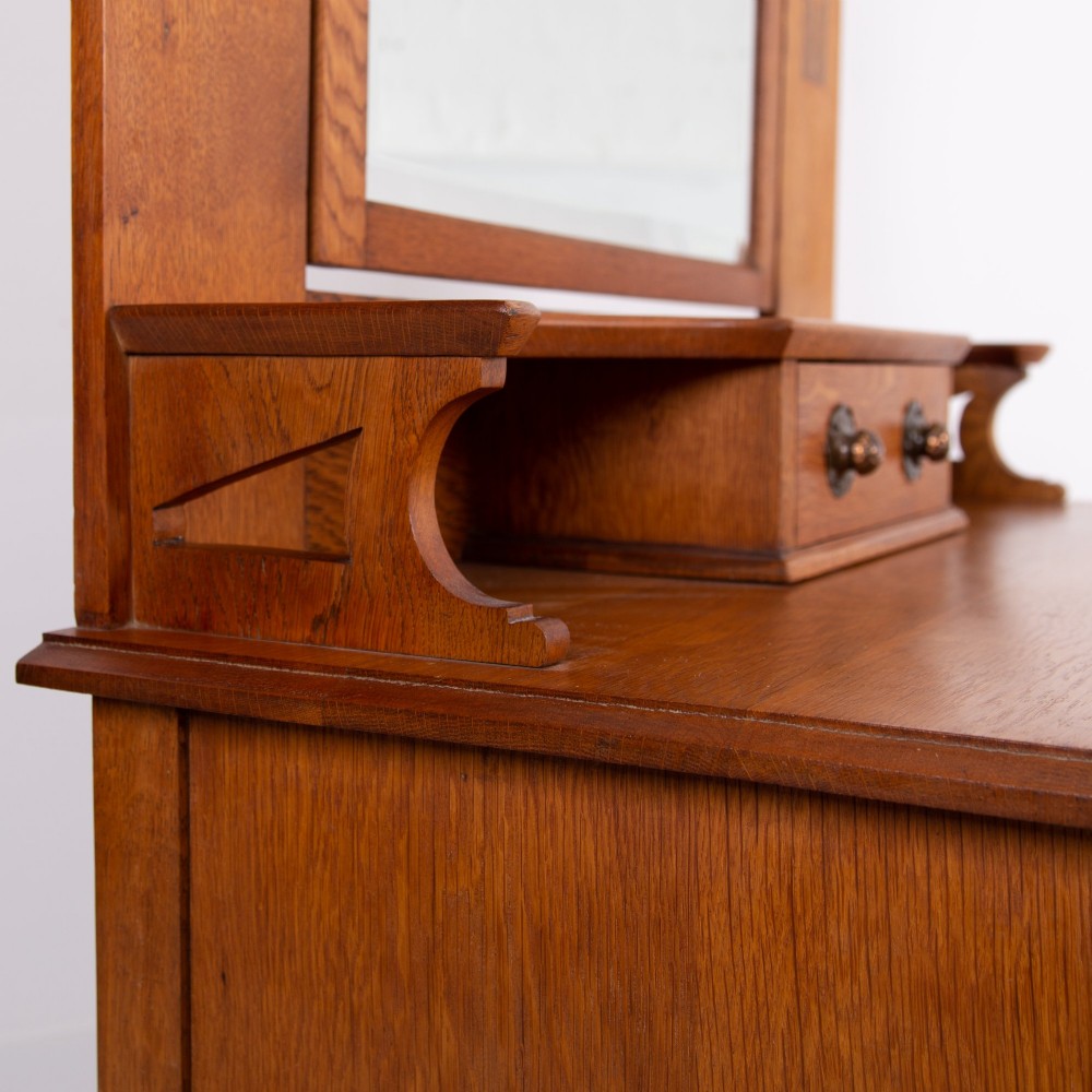 An Arts and Crafts oak dressing chest featuring a superstructure with a central mirror, inlaid side panels, and small jewellery drawers above a chest of drawers. View XII