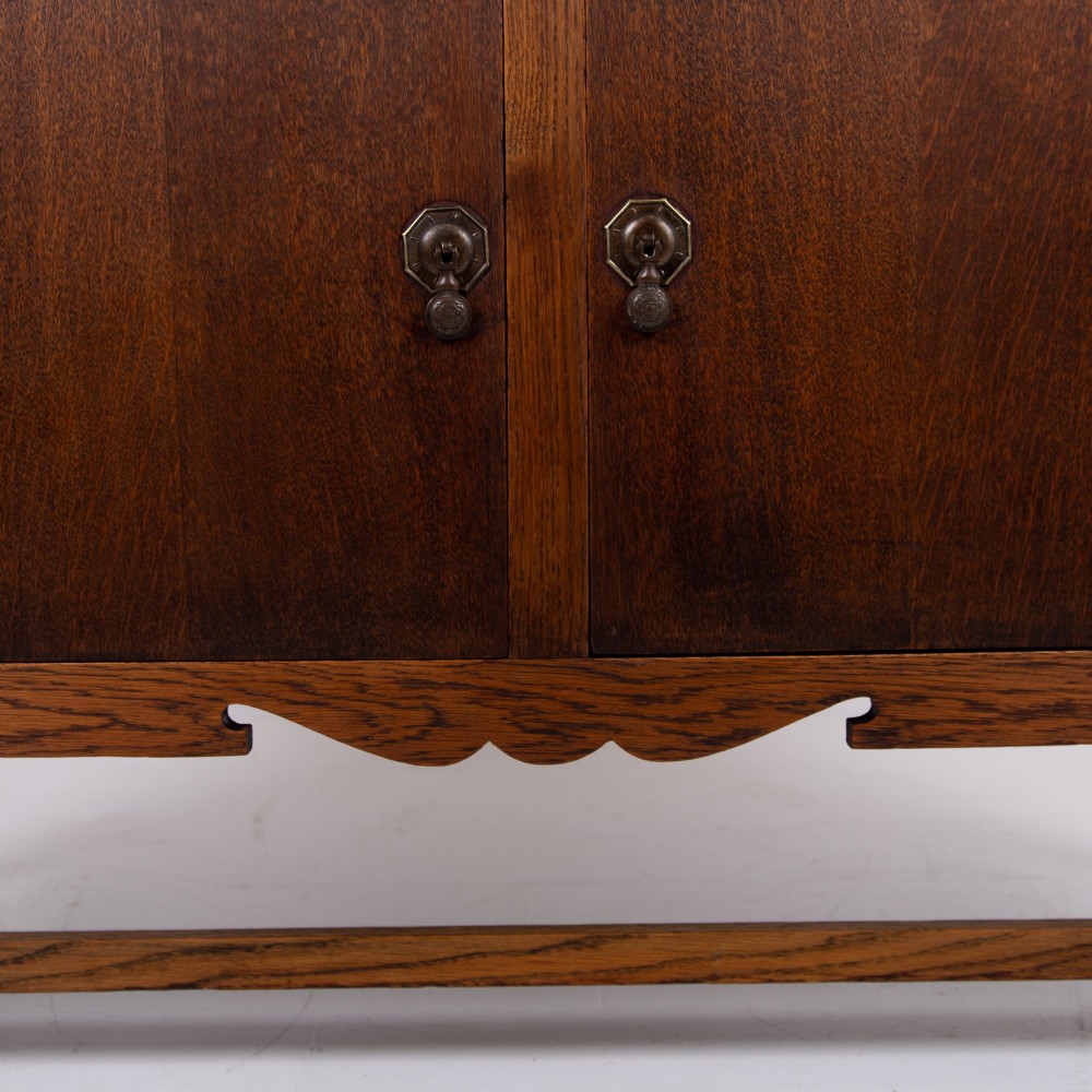 A 1930s warm brown oak sideboard featuring two carved top drawers above a double-door cupboard, supported by block legs and a low stretcher base. View VI