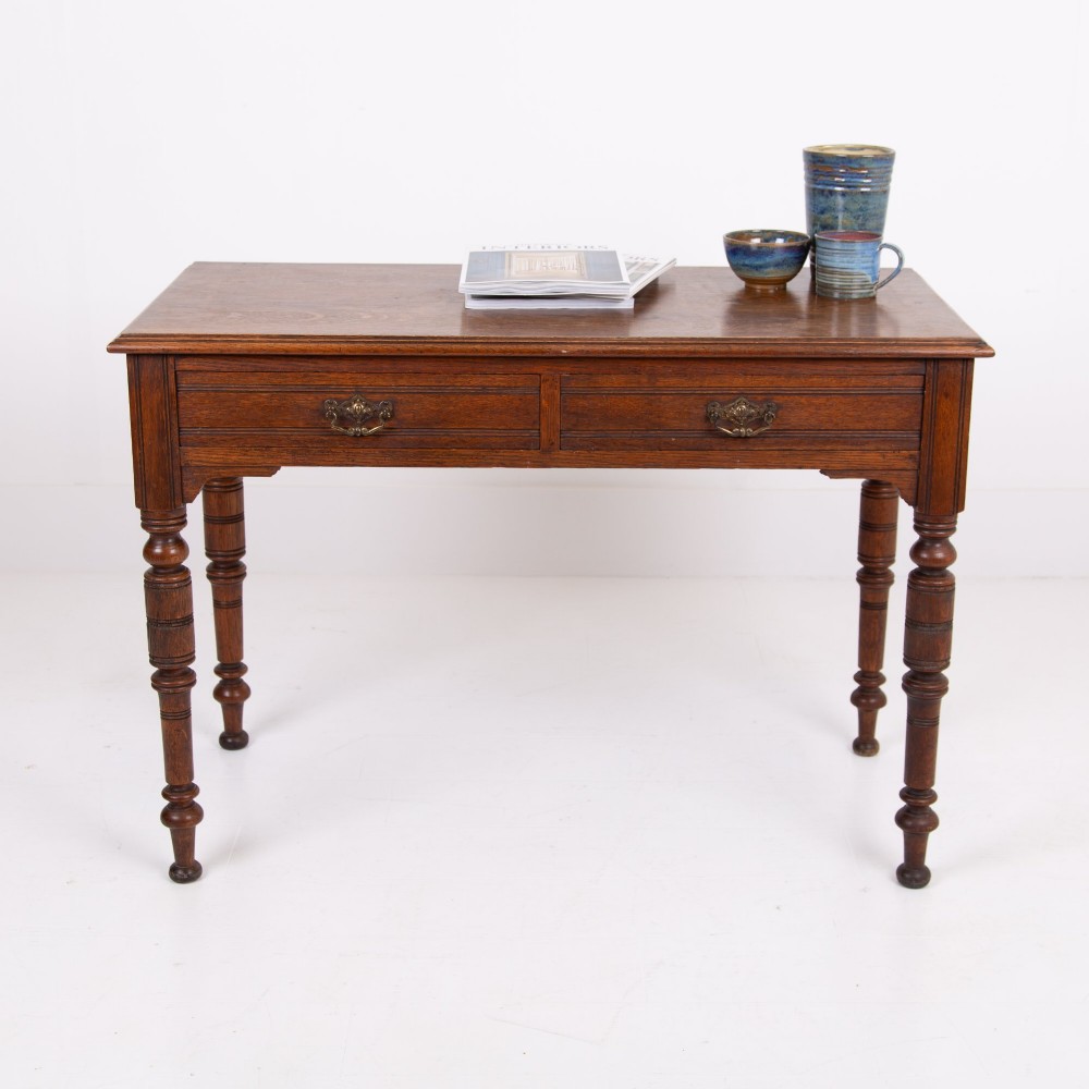 A Victorian solid oak writing desk featuring two drawers with brass drop handles and supported by four turned legs.