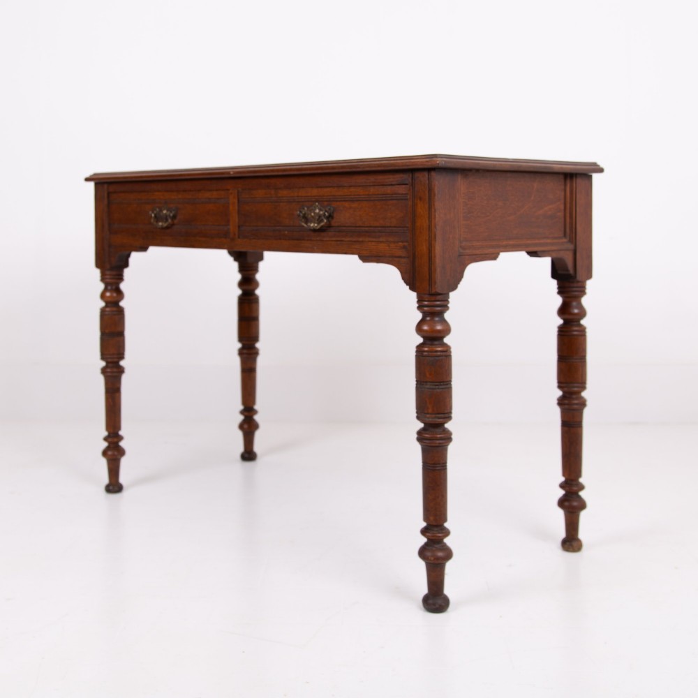 A Victorian solid oak writing desk featuring two drawers with brass drop handles and supported by four turned legs. View VII
