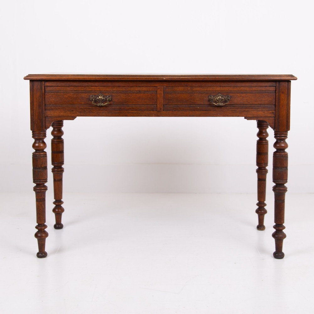 A Victorian solid oak writing desk featuring two drawers with brass drop handles and supported by four turned legs. View XII