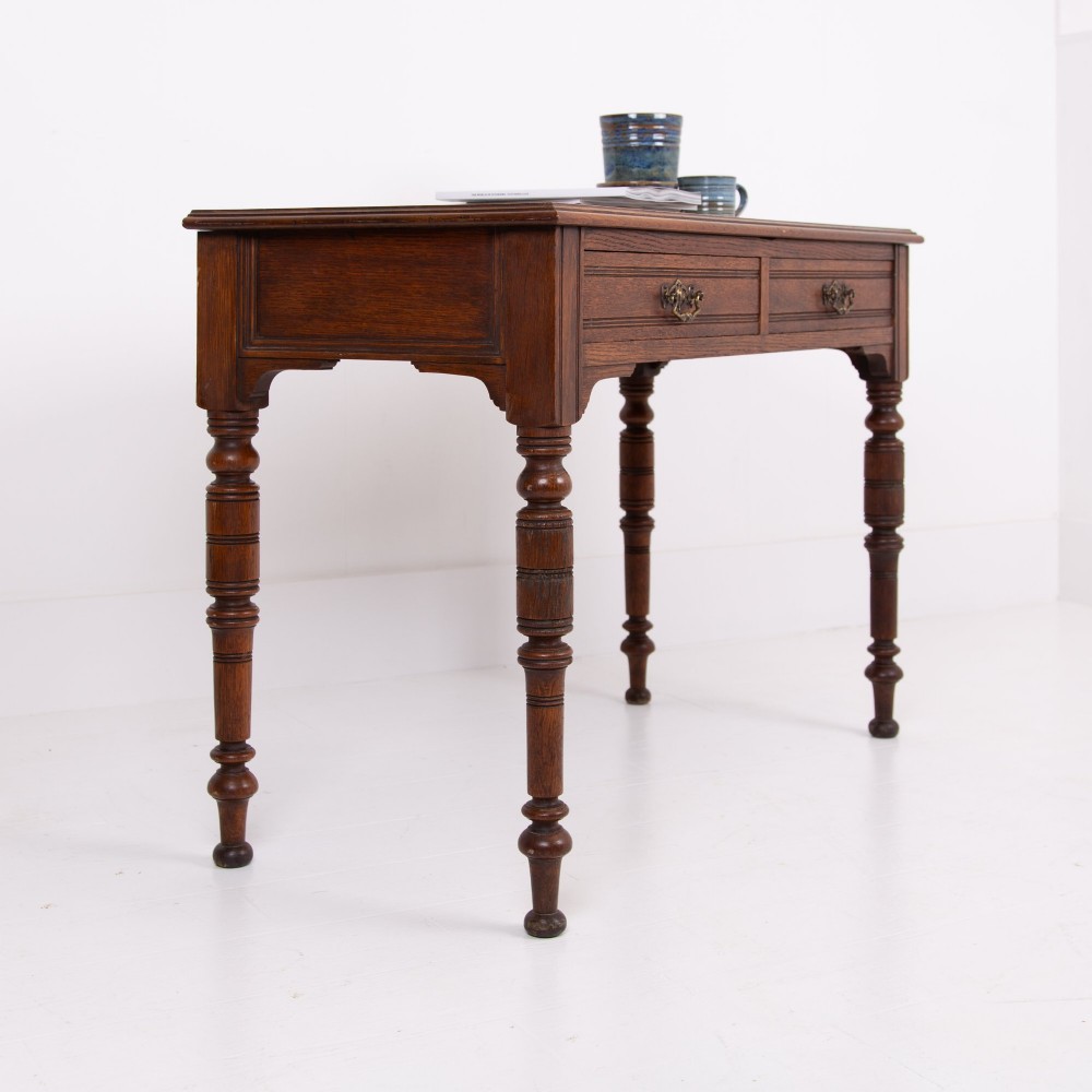 A Victorian solid oak writing desk featuring two drawers with brass drop handles and supported by four turned legs. View XIII