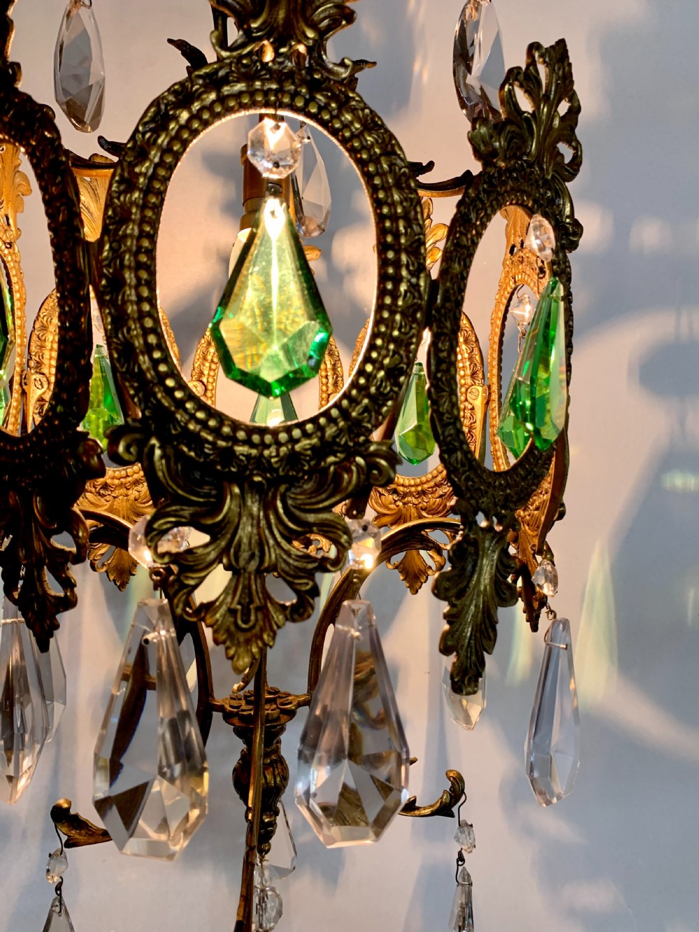 An ornate gilded bronze Florentine pendant light featuring scrolling leaf motifs, oval frame inserts enclosing faceted green crystals, and hanging clear glass prisms. View II