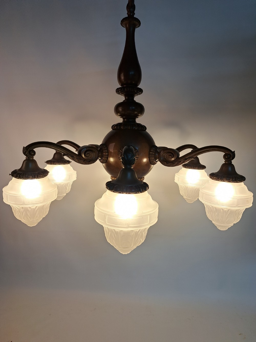A five-arm dark bronze chandelier featuring a central turned stem, curved scroll arms, and downward-facing frosted glass shades.