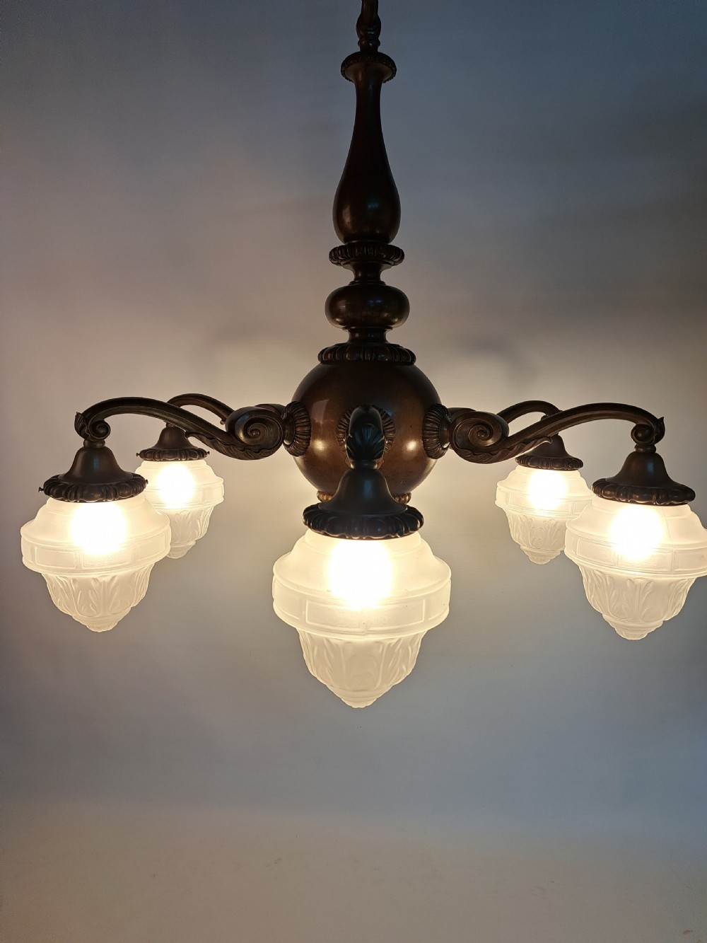 A five-arm dark bronze chandelier featuring a central turned stem, curved scroll arms, and downward-facing frosted glass shades. View II