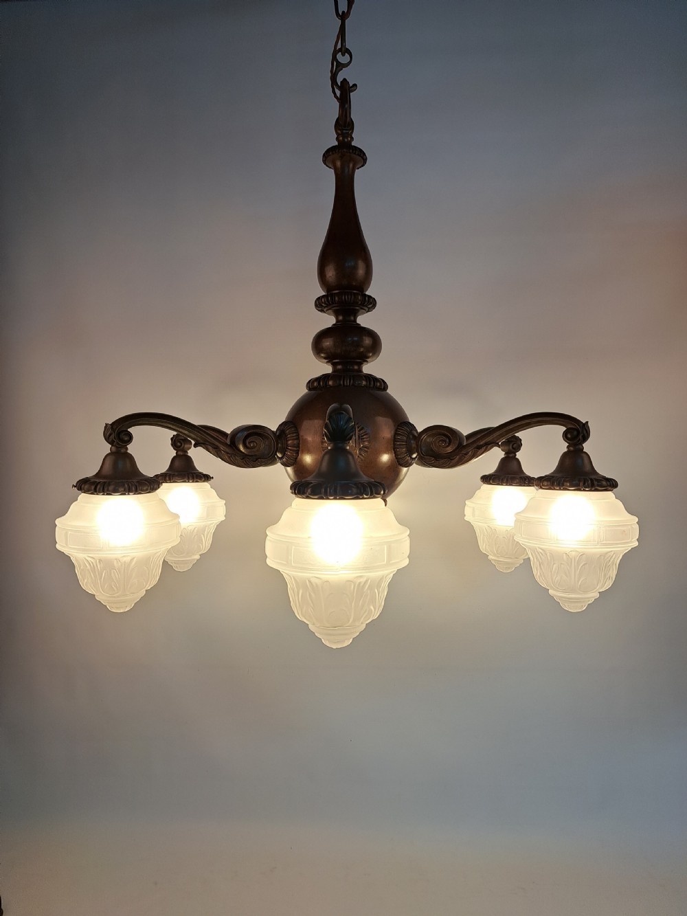 A five-arm dark bronze chandelier featuring a central turned stem, curved scroll arms, and downward-facing frosted glass shades. View III