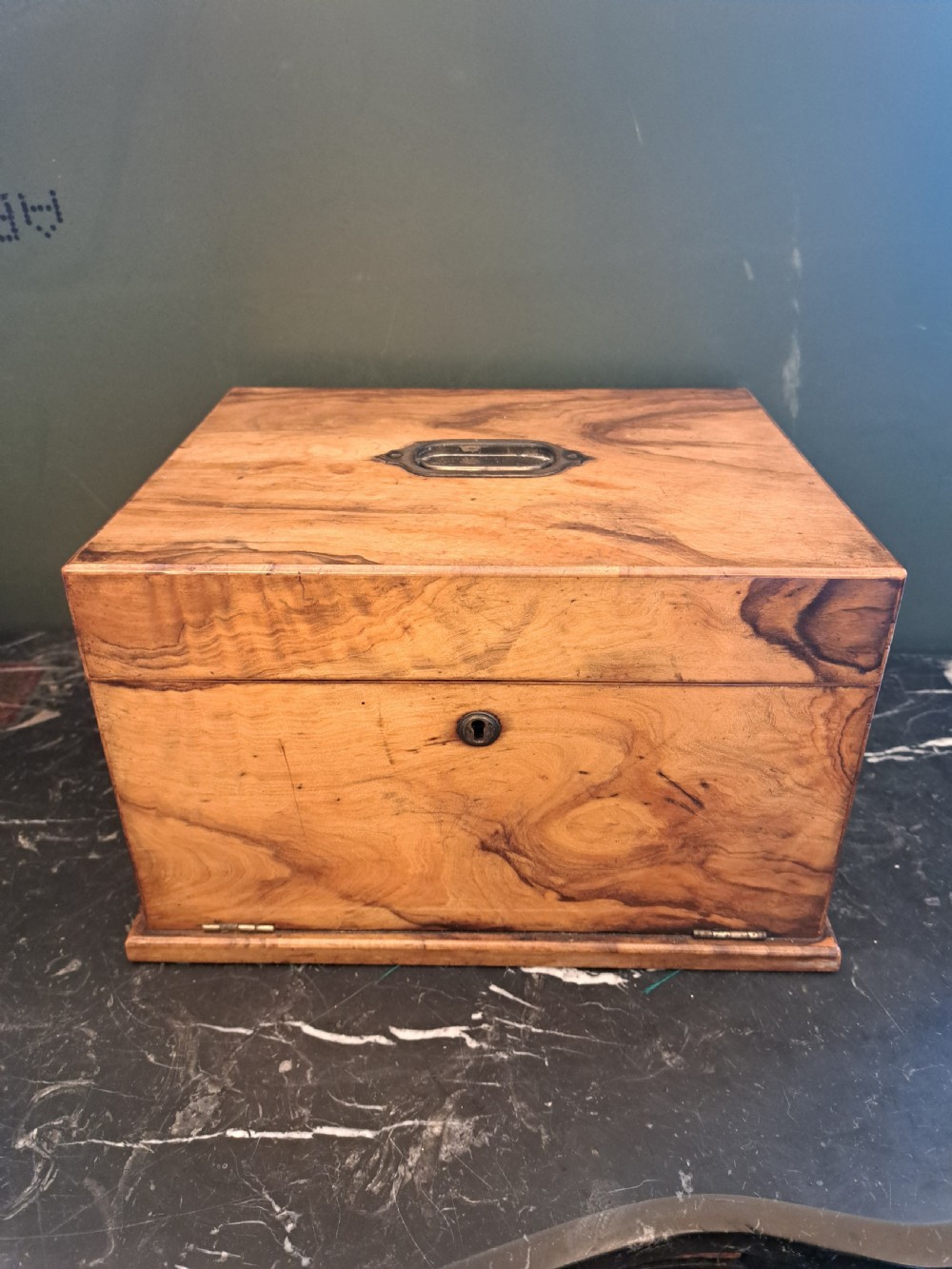 A rectangular blonde walnut jewellery box with a brass keyhole escutcheon and recessed handle on the lid, resting on a dark marble surface.