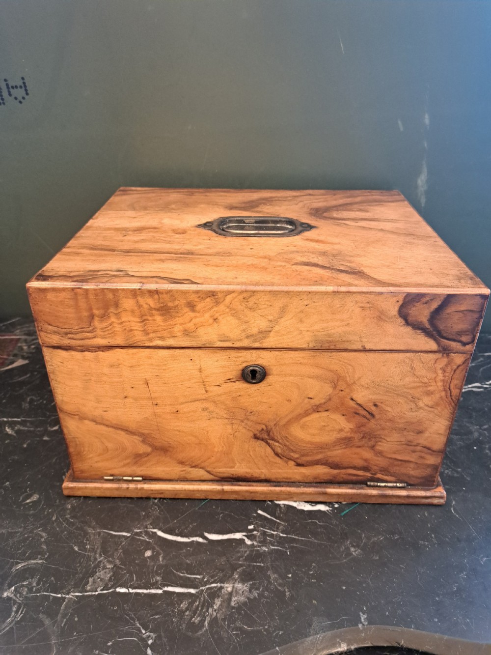 A rectangular blonde walnut jewellery box with a brass keyhole escutcheon and recessed handle on the lid, resting on a dark marble surface. View IV