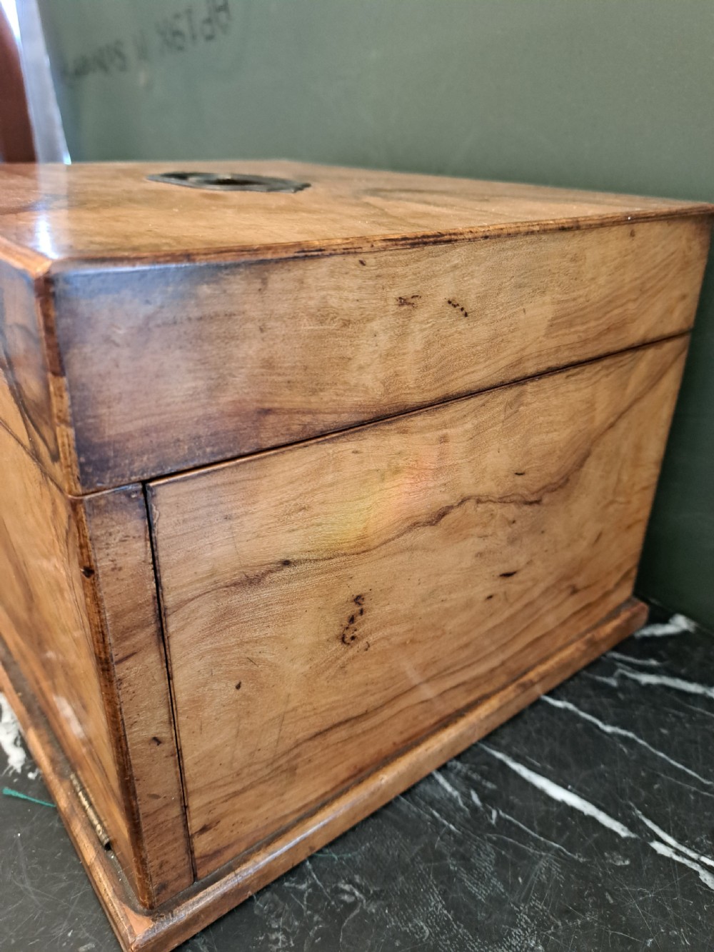 A rectangular blonde walnut jewellery box with a brass keyhole escutcheon and recessed handle on the lid, resting on a dark marble surface. View V