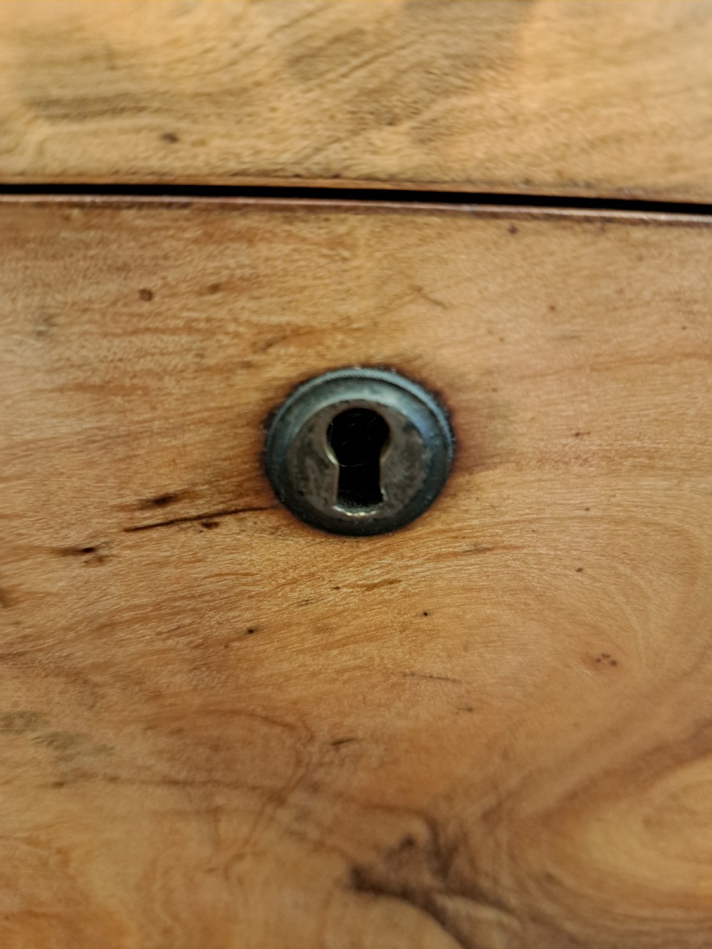 A rectangular blonde walnut jewellery box with a brass keyhole escutcheon and recessed handle on the lid, resting on a dark marble surface. View XI