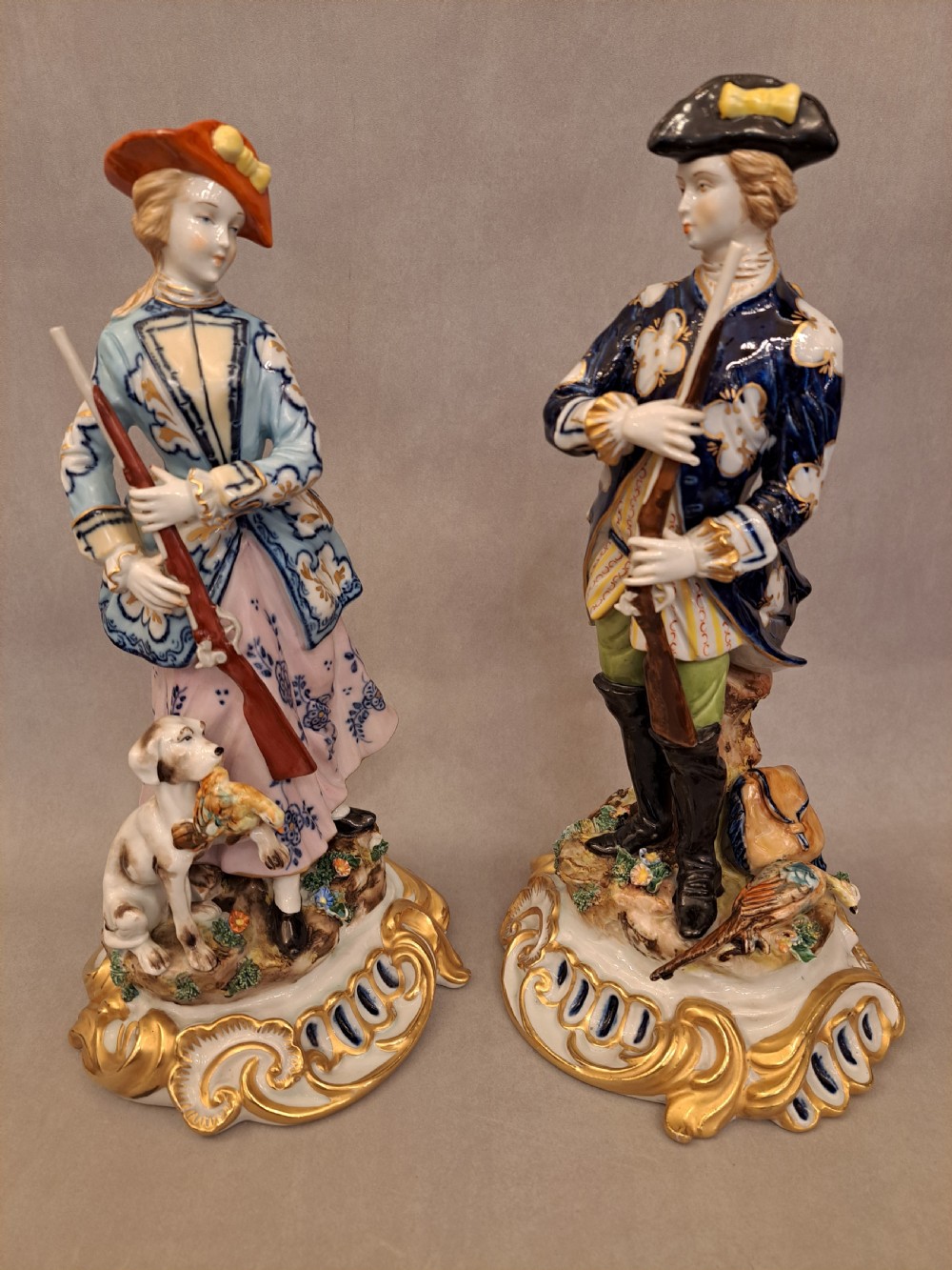 A pair of hand-painted Capodimonte porcelain figurines depicting a male and female hunter in 18th-century clothing holding muskets, positioned on gilt scrollwork bases.