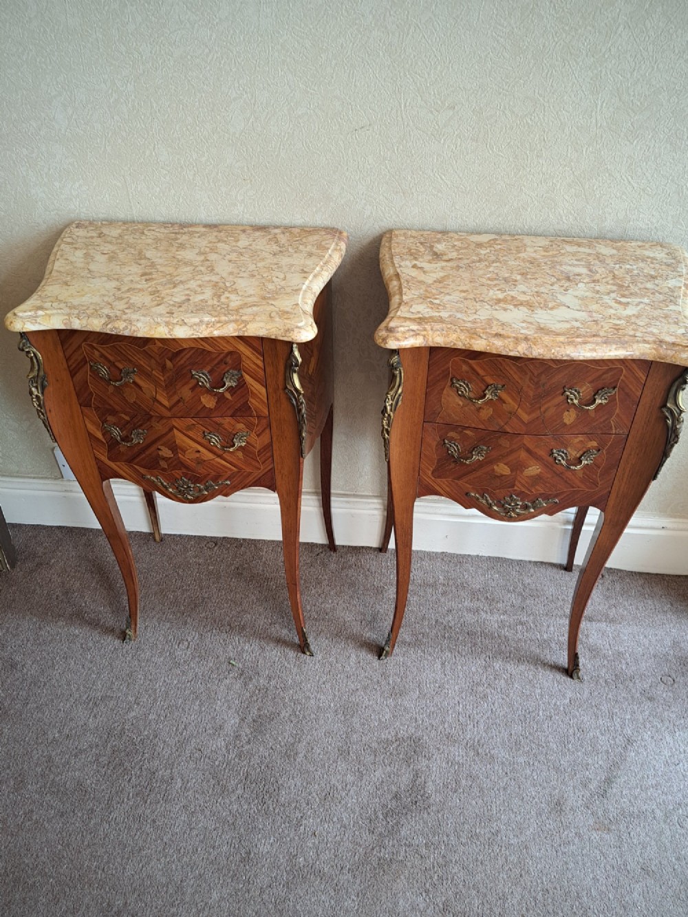 A pair of French style bedside tables featuring contoured beige marble tops, floral marquetry veneer drawers, brass handles, and sweeping cabriole legs.