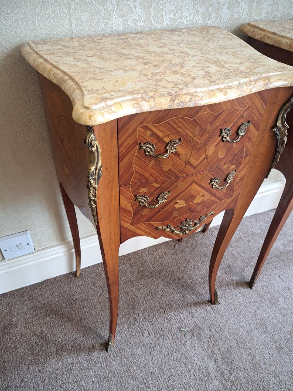 A pair of French style bedside tables featuring contoured beige marble tops, floral marquetry veneer drawers, brass handles, and sweeping cabriole legs. View X