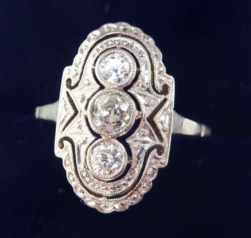 fig 1. An Art Deco 18ct white gold ring featuring three vertically aligned round diamonds within an oval pierced filigree plaque with millgrained borders.