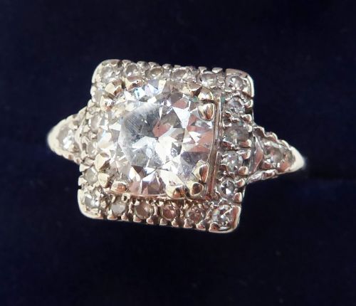 fig 1. An Art Deco style 18ct white gold ring featuring a central diamond enclosed in a square geometric halo setting with embellished shoulders.