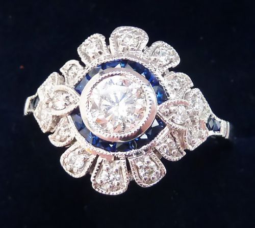 fig 1. An Art Deco white gold target ring featuring a central round diamond encircled by calibre-cut blue sapphires and a outer scalloped diamond halo with milgrain borders.