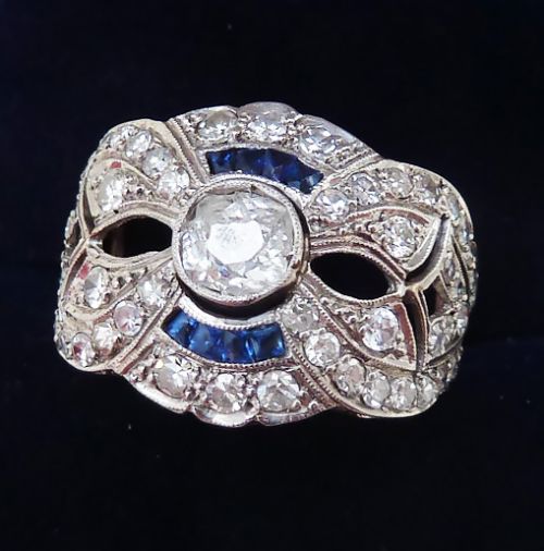 fig 1. An Art Deco platinum cluster ring featuring a central round diamond surrounded by blue sapphire arcs, geometric openwork, and smaller accent diamonds.